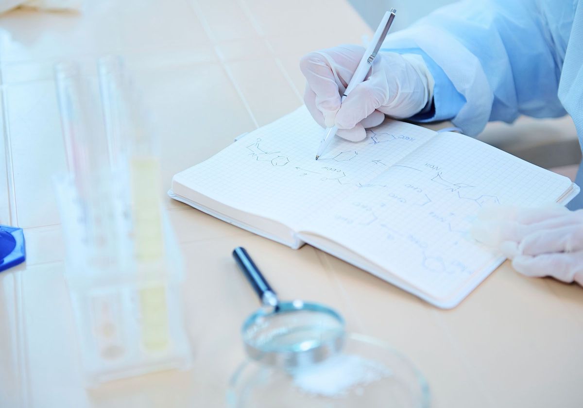 A scientist with gloved hands sitting at a table with a magnifying glass and lab glassware and writing in a notepad with a pen. A scientist with gloved hands sitting at a table with a magnifying glass and lab glassware and writing in a notepad with a pen.