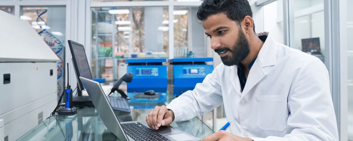 A person sitting at a laboratory bench, typing on a laptop while looking at notes on a clipboard. A person sitting at a laboratory bench, typing on a laptop while looking at notes on a clipboard.