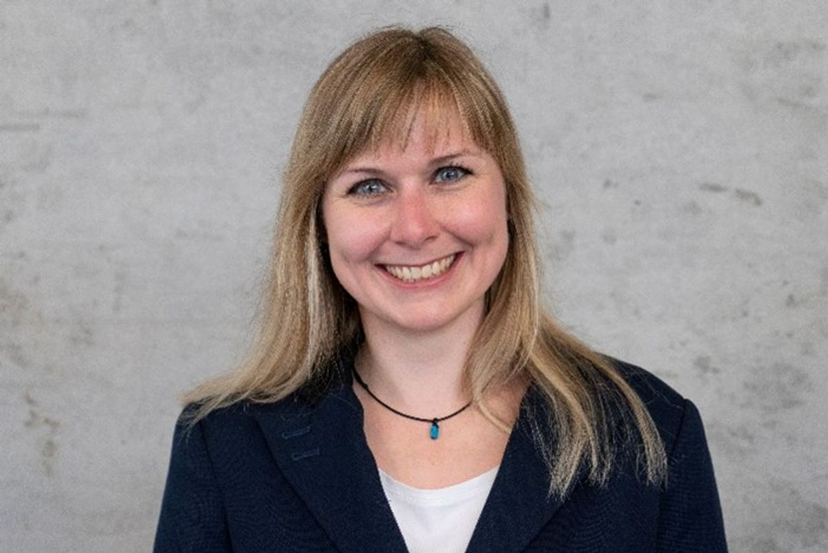 Scientist wearing a black blazer and white shirt smiling to the camera with a light grey wall in the background. Scientist wearing a black blazer and white shirt smiling to the camera with a light grey wall in the background.