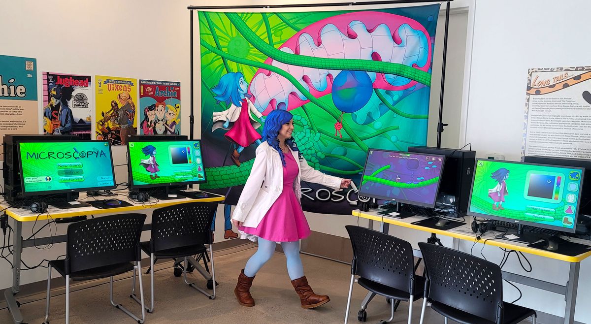 Beata Mierzwa, dressed in a pink dress, lab coat, and sporting blue hair, resembles the animated character from the game, Microscopya, she and her team designed. Computer stations beside her have the game pulled up at the 2022 San Diego Comic-Con. Beata Mierzwa, dressed in a pink dress, lab coat, and sporting blue hair, resembles the animated character from the game, Microscopya, she and her team designed. Computer stations beside her have the game pulled up at the 2022 San Diego Comic-Con.