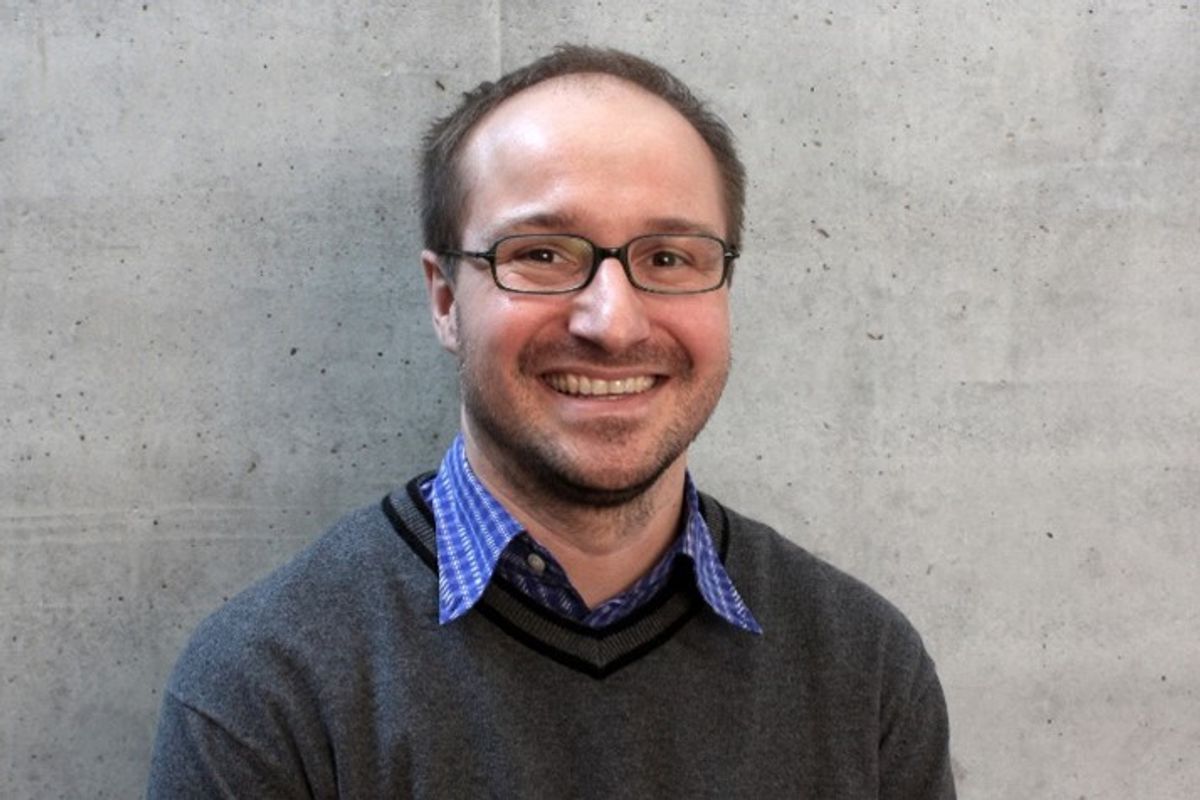 Scientist wearing a dark grey sweater, blue checkered shirt, and glasses smiling to the camera with a light grey wall in the background. Scientist wearing a dark grey sweater, blue checkered shirt, and glasses smiling to the camera with a light grey wall in the background.