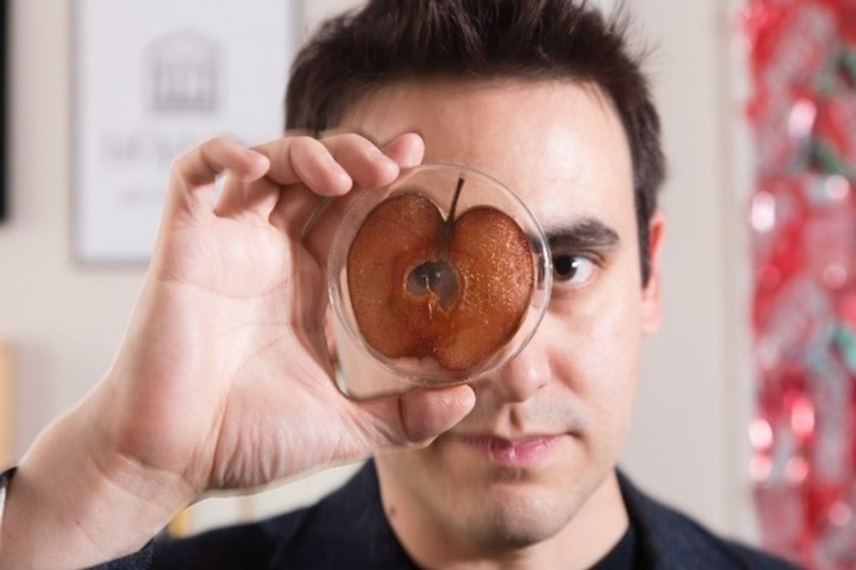 A photo of Andrew Pelling holding a Petri dish with a browning apple inside of it close to his face. A photo of Andrew Pelling holding a Petri dish with a browning apple inside of it close to his face.