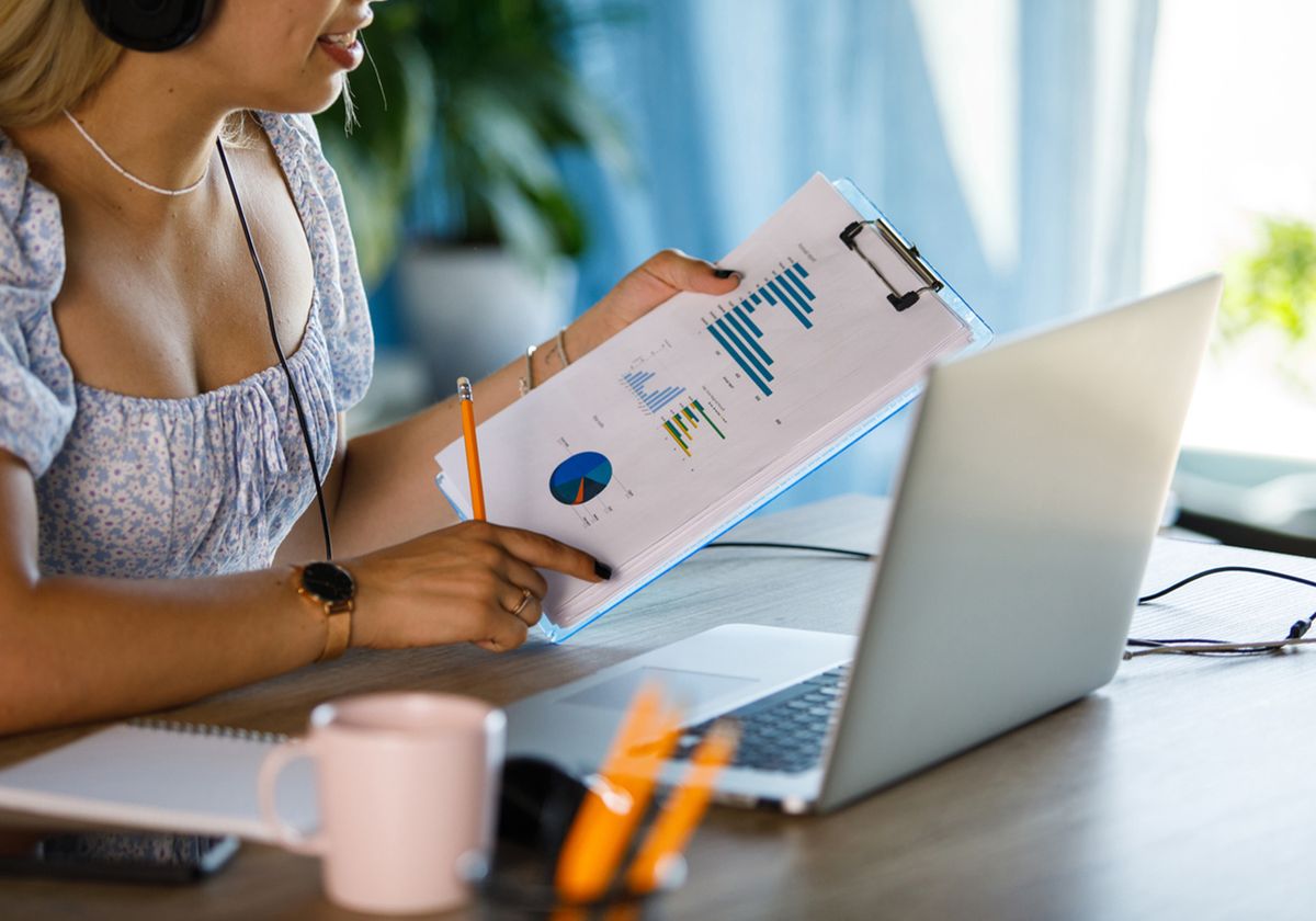 An individual looking at graphs and charts on a clipboard in front of a laptop. An individual looking at graphs and charts on a clipboard in front of a laptop.