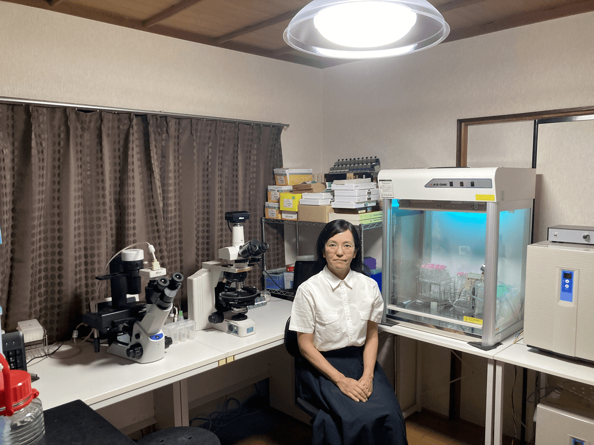 The researcher, Kyoko Hagino sits next to two microscopes, sterile hood, curtains, and she wears a blue skirt and white blouse. The researcher, Kyoko Hagino sits next to two microscopes, sterile hood, curtains, and she wears a blue skirt and white blouse.