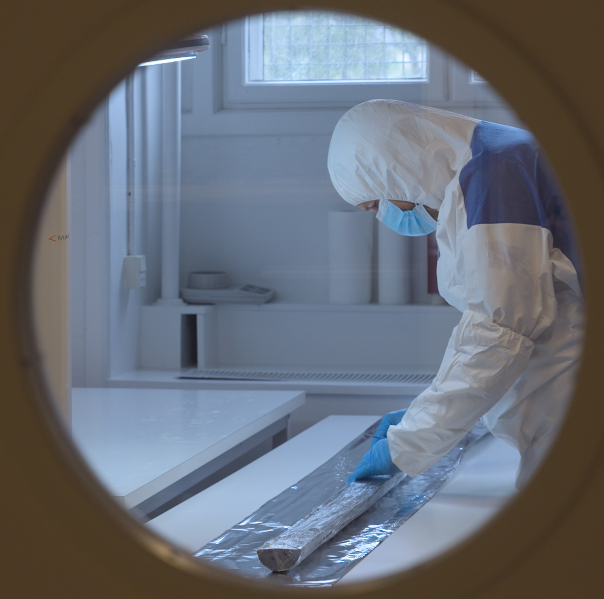 A photo through a door of a researcher in PPE working with a sediment core A photo through a door of a researcher in PPE working with a sediment core